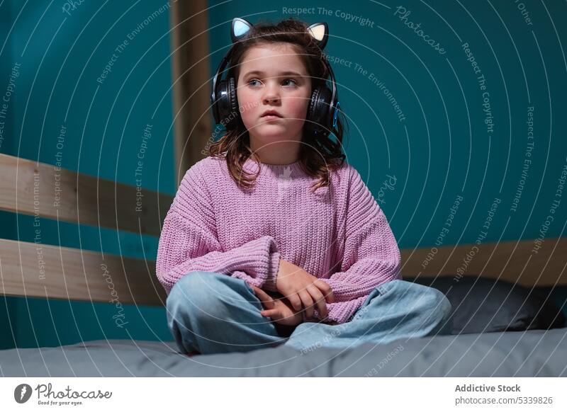 Calm child in headphones sitting on bed kid music listen legs crossed adorable thoughtful bedroom gadget little home device comfort at home calm cozy inside
