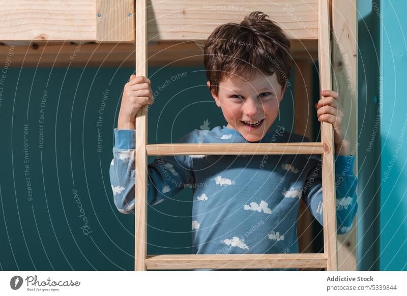 Cheerful boy behind ladder of bunk bed in bedroom pajama bed time fun portrait smile cute happy kid childhood cheerful wooden joy casual style optimist content