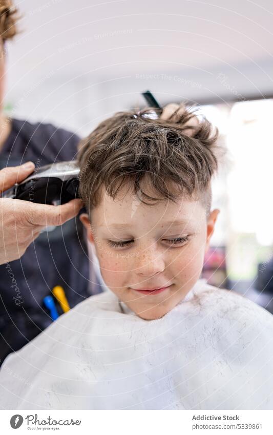 Anonymous stylist cutting hair of child client kid chair hairstyle haircut fabric studio comfort calm comb salon care machine equipment childhood customer