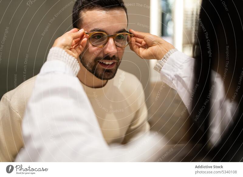 Anonymous female eye doctor helping male customer to wear eyeglasses man eyewear choose select store optical smile ophthalmologist eyesight vision shop