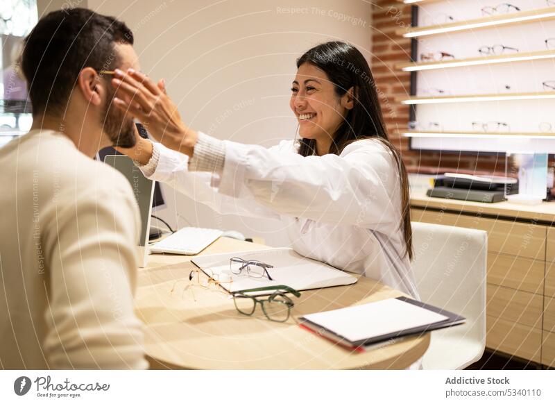 Cheerful female eye doctor helping male customer to wear eyeglasses man eyewear choose select store optical smile ophthalmologist eyesight vision shop