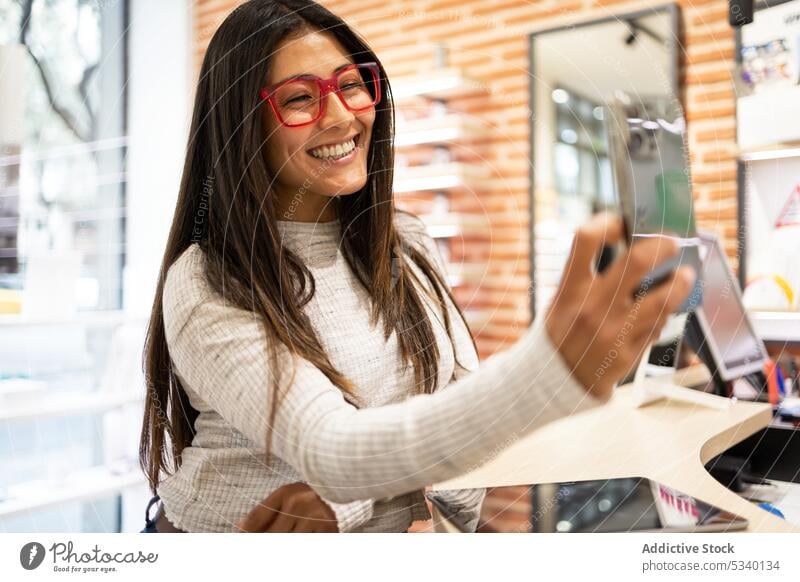 Happy female in eyewear taking selfie in optical shop woman customer buy eyeglasses smartphone using shelf choose cheerful happy store mobile smile gadget