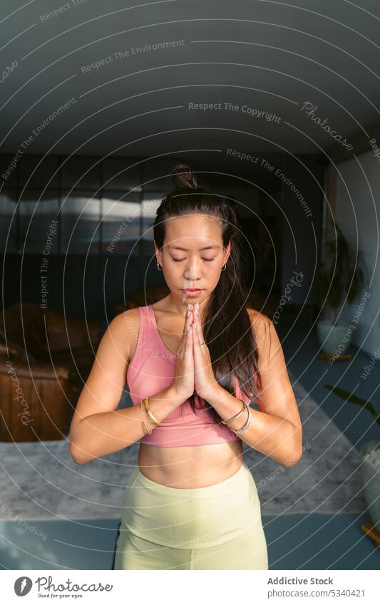 Calm Asian woman with clasped hands in living room yoga namaste meditate home practice mindfulness tranquil eyes closed asana calm harmony female ethnic asian