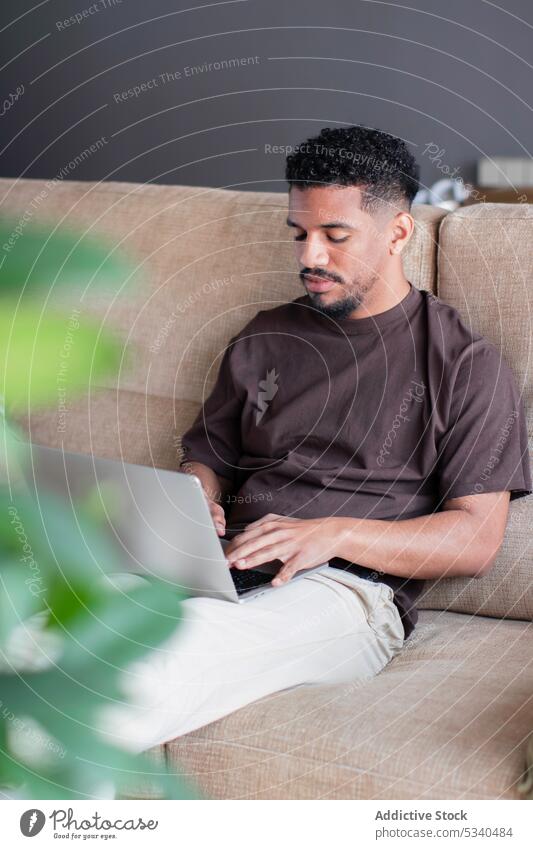 Serious ethnic man working on laptop at home freelance using typing online remote internet concentrate young sofa african american casual gadget telework