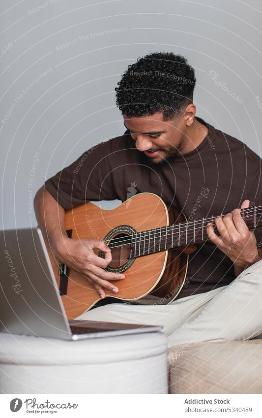 Cheerful ethnic man playing guitar with laptop on sofa using positive online music acoustic guitarist musician instrument hobby young african american melody