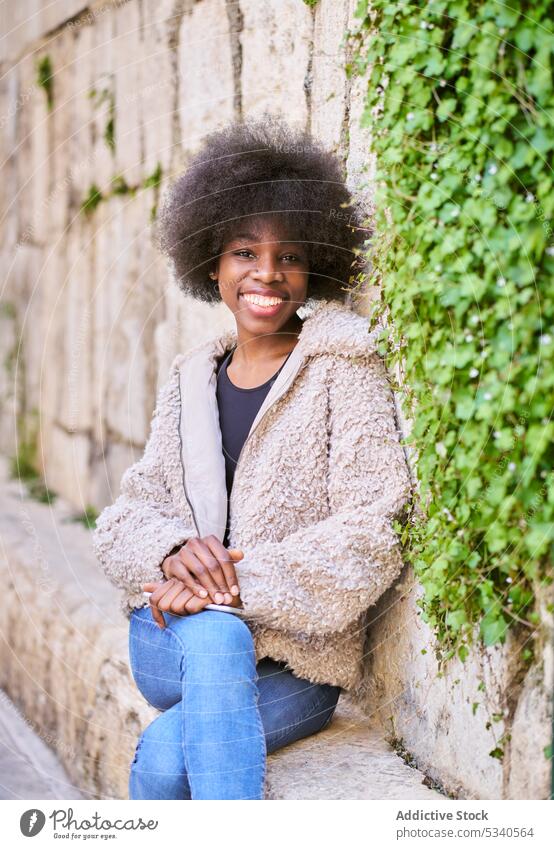 Cheerful black teen with Afro hairdo sitting on concrete terrace by stone wall coat happy urban positive cheerful smile joy african american female content