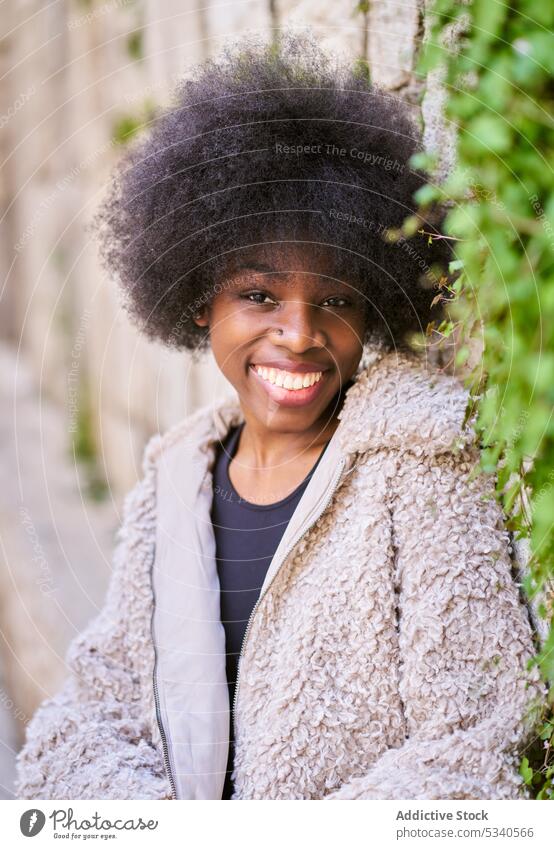 Cheerful black teen with Afro hairdo on concrete terrace by stone wall coat happy urban positive cheerful smile joy african american female content young