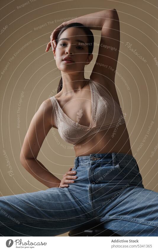 Graceful woman sitting on chair in light studio style lingerie portrait model thoughtful sensual stool jeans fashion underwear figure arms raised female