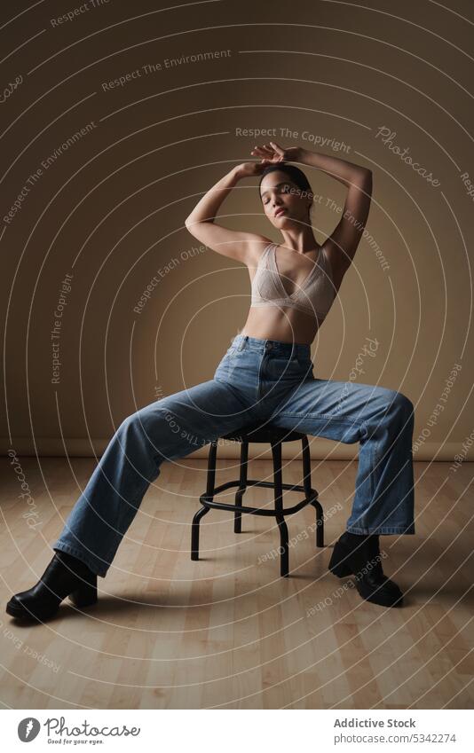Graceful woman sitting on chair in light studio style lingerie portrait model thoughtful sensual stool jeans fashion eyes closed underwear figure arms raised