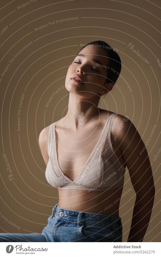 Dreamy woman in bra sitting with closed eyes against beige background model calm hand behind back eyes closed portrait style individuality studio jeans female