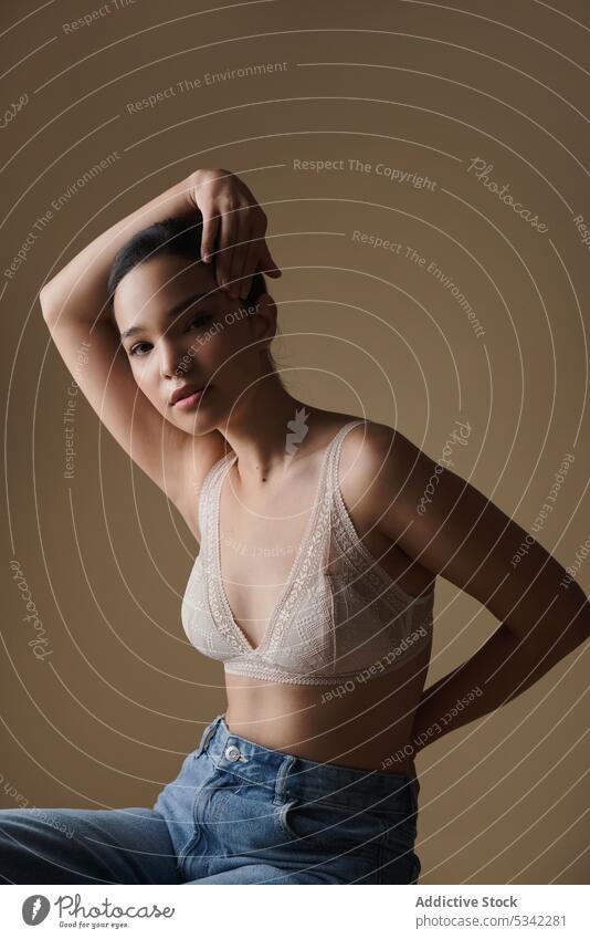 Alluring woman with hand on head and behind back in studio model bra hand behind back stool confident jeans style portrait feminine trendy fashion female young