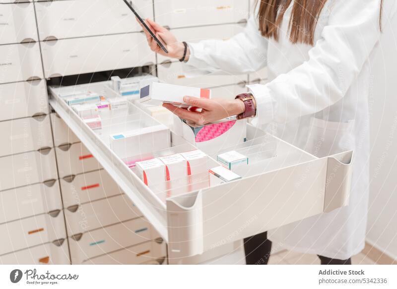 Crop pharmacist using tablet while standing by slotted shelves with drugs in modern pharmacy scientist laboratory woman drugstore device gadget medication shop