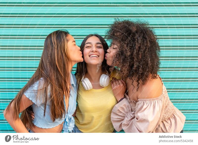 Happy diverse women kissing friend cheeks girlfriend together smile friendship cheerful best friend relationship multiracial multiethnic young street bonding
