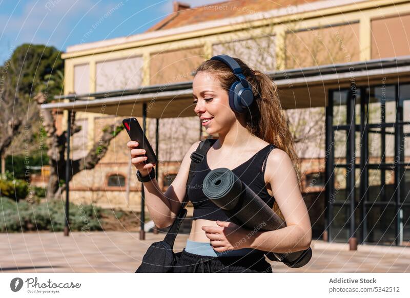 Smiling woman in headphones using smartphone listen music street walk mat browsing activity carry device female young surfing online relax social media internet