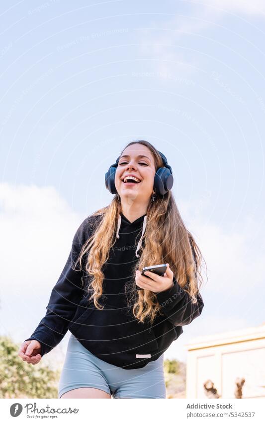 Smiling woman with smartphone listening to music in headphones using cheerful joy enjoy carefree gadget wireless happy device female young smile casual blue sky