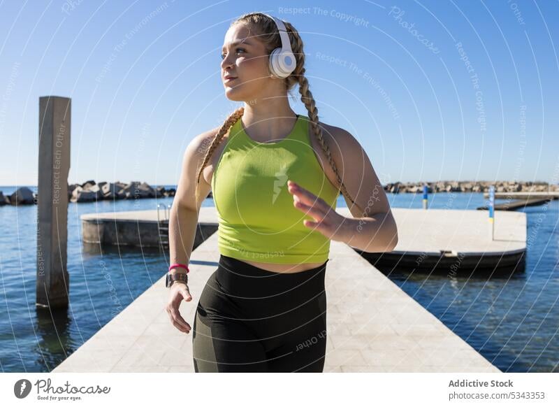 Fit sportswoman with headphones jogging on pier athlete training run fitness workout exercise healthy water wellness sportswear listen cardio sun female