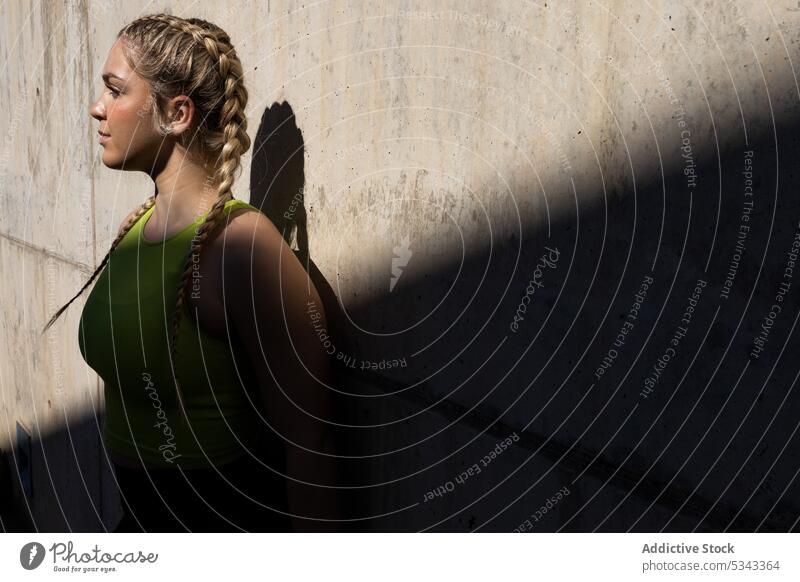 Young woman standing near concrete wall with shadow sportswoman portrait confident athlete sportswear street sunlight young female dreamy pensive serious sporty