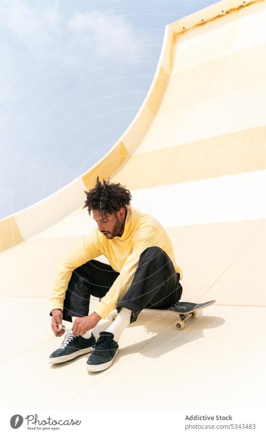 Stylish black man sitting on skateboard over skate ramp in daylight skater skate park shoelace tie sneakers activity blue sky energy hobby extreme ethnic male