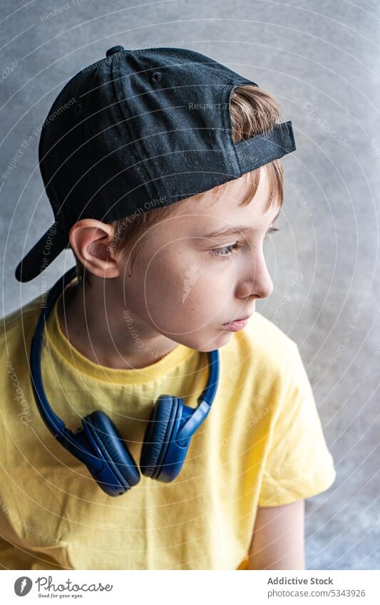 Side view of pensive kid in yellow t-shirt and black cap with headphones on the neck looking away against blurred background beautiful boy child ear eyes face