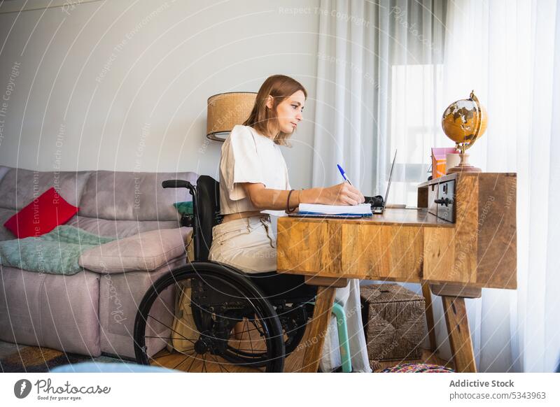 Young woman sitting on wheelchair while taking notes from laptop notebook take note write freelance sofa living room home using gadget at home planner work