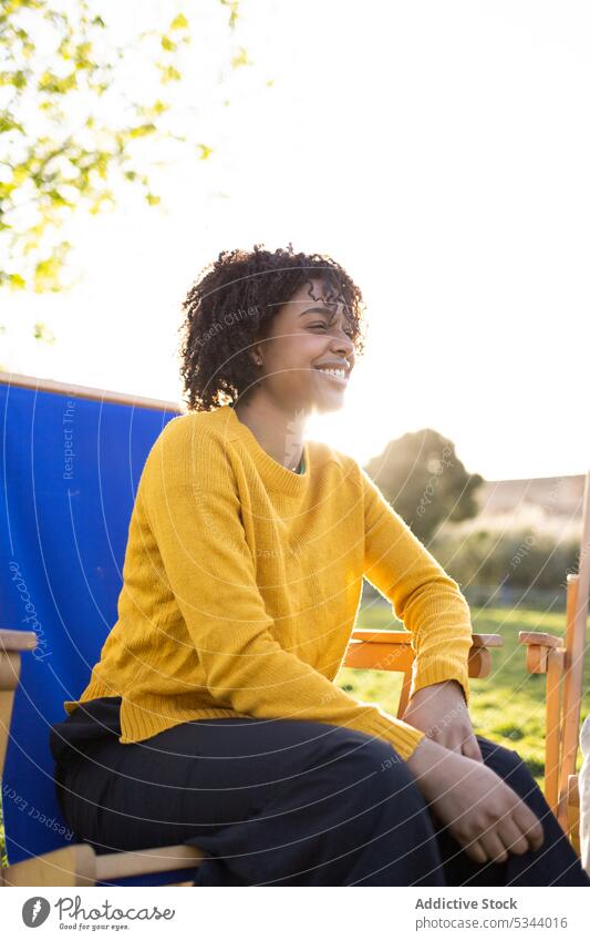 Cheerful young ethnic woman sitting on chair in park nature tree content glad positive smile enjoy summer happy hand on cheek female african american black