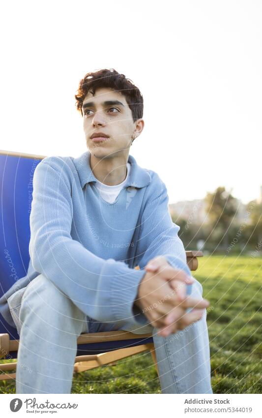 Thoughtful young man sitting on chair in park relax ponder hands clasped summer pensive thoughtful calm sunlight male casual think weekend daytime nature