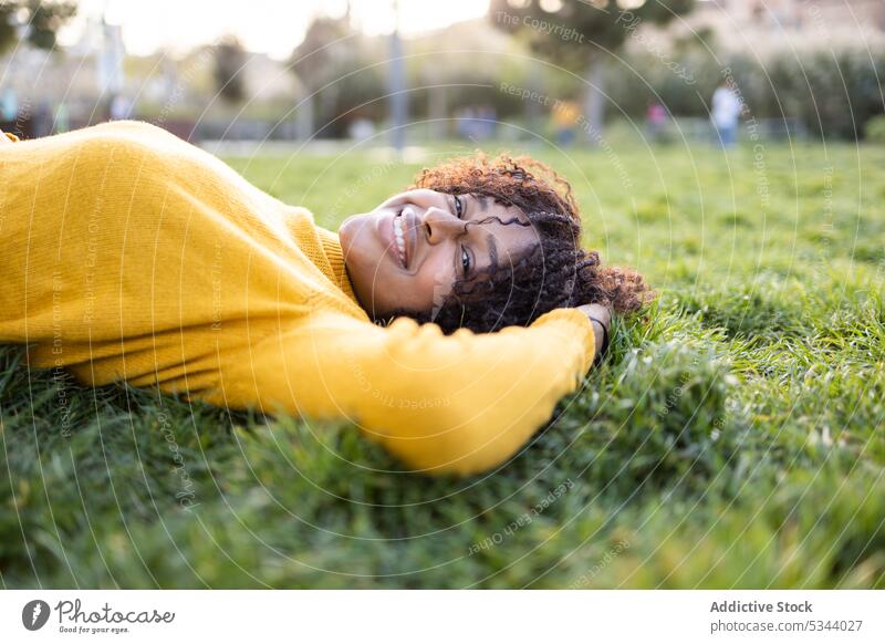 Happy ethnic woman with curly hair lying on grass smile park nature tree lawn green content positive female young african american black happy sweater daytime
