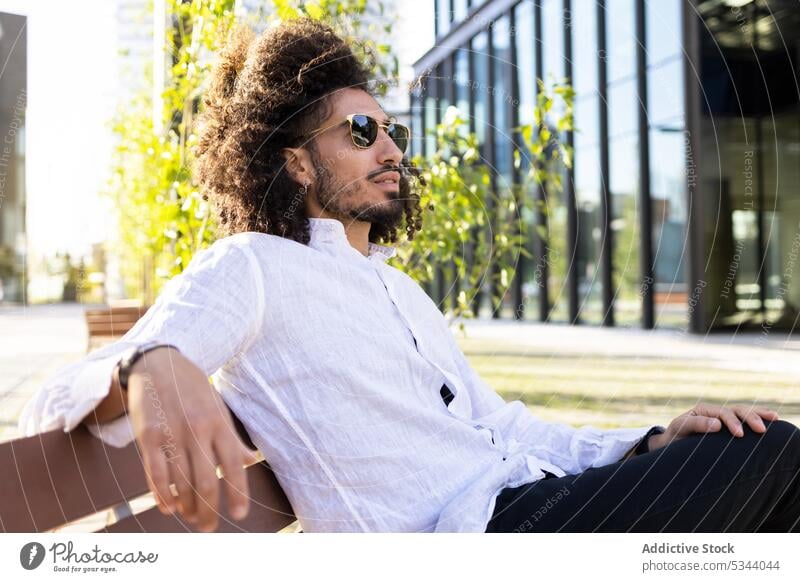 Stylish ethnic man with curly hair sitting on bench trendy style park confident afro serious appearance sunglasses male african american outfit black man cool