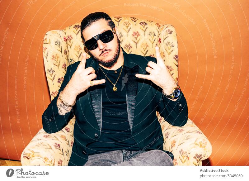 Calm man showing rock gestures in armchair in studio style sunglasses hipster serious confident accessory trendy male fashion beard cool young jacket