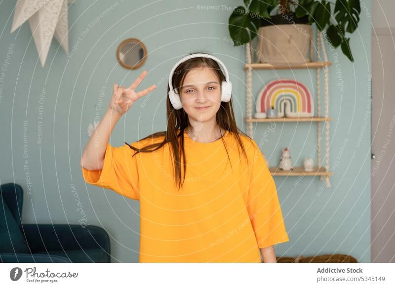 Cheerful child showing peace sign girl music listen v sign dance gesture headphones bedroom wireless using positive female device home happy demonstrate smile