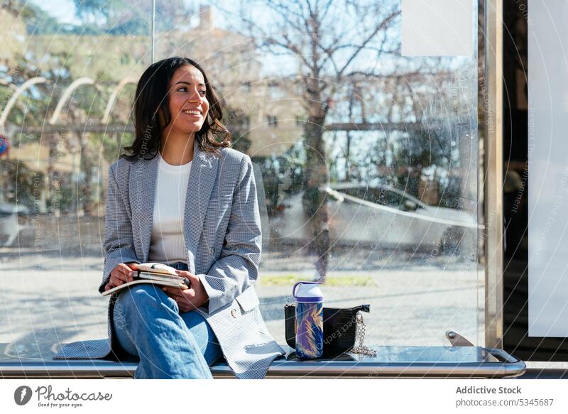 Smiling ethnic woman sitting on bench in city bus stop student textbook smile street water glass wall female young positive study legs crossed content daylight