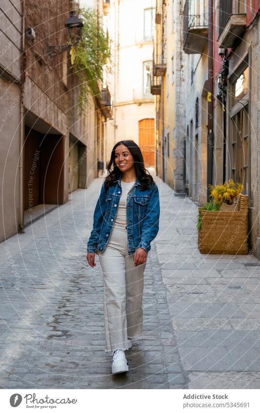Cheerful ethnic woman walking on city street alley town stroll shadow building exterior narrow daytime young pavement summer urban residential female hispanic
