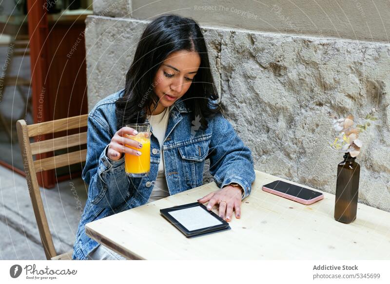 Concentrated ethnic woman holding a glass of orange juice in a street cafe using tablet drink internet online smartphone cellphone young social media message