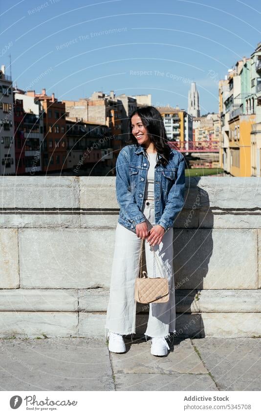 Happy ethnic woman standing near wall on terrace smile handbag blue sky building urban glad cityscape cloudless happy young summer satisfied female hispanic