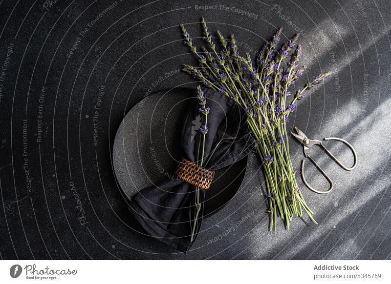 High angle of summer table setting with lavender flowers on black concrete dark background between a napkin on plate and scissors bouquet cement concept cutlery