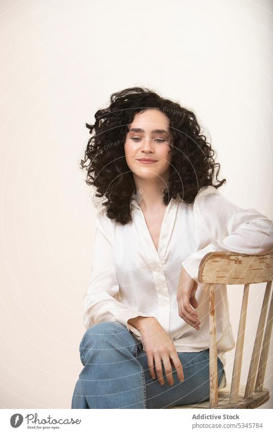 Young woman sitting on wooden chair in studio legs crossed curly hair portrait style confident casual appearance female young jeans model calm happy outfit