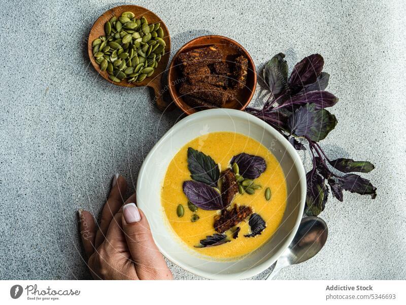 Cropped hand touching bowl with pumpkin cream soup with basil herb on table woman colorful creamy crusty detox diet dinner dinnerware eat food fresh healthy
