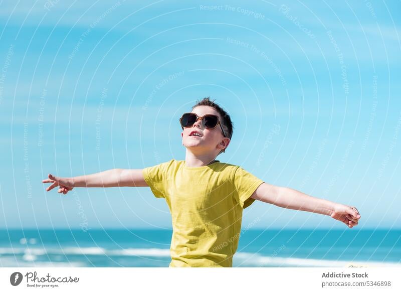 Boy with outstretched arms enjoying wind on beach boy sea water freedom blue sky child vacation happy sun glad childhood positive optimist sunglasses sunlight