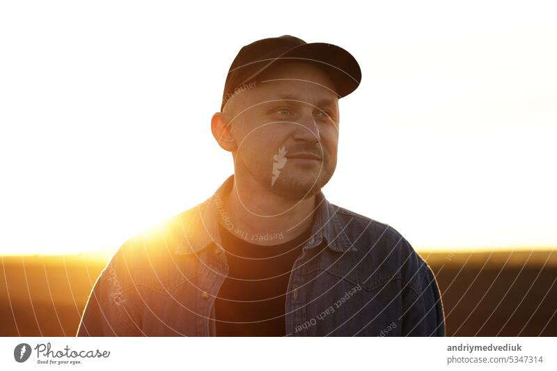 Portrait of smiling male farmer in cap is walking through endless plowed farmland field on sunset. Professional agronomist inspecting cultivated land in sunlight. Agribusiness concept and farming.