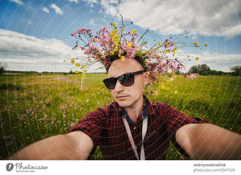 Young smiling man in a wildflower wreath and sunglasses takes a selfie on a wide-angle camera is having fun in the field on summer day. Positive guy doesn't have allergy. Lifestyle summer Concept.