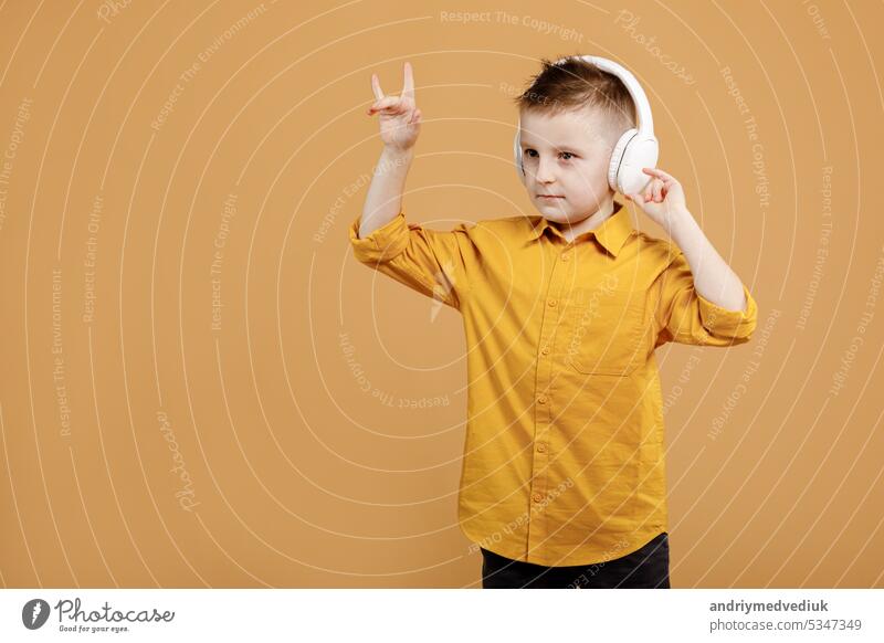Portrait of funny clever school boy with headphones in yellow shirt. Yellow studio background. Education. Looking, smiling and shows a thumb up at camera. High quality photo