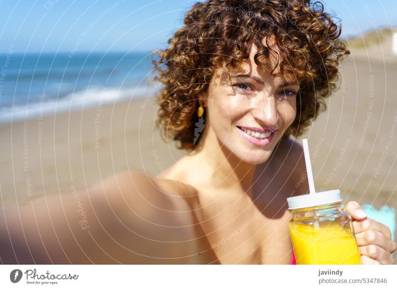 Cheerful woman enjoying juice and taking self portrait on beach selfie drink summer vacation smile refreshment carefree cheerful happy beverage curly hair young