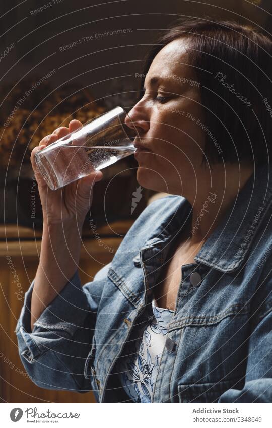 Calm mature woman drinking water from glass kitchen peaceful eyes closed calm beverage thirst home refreshment female tranquil jacket short hair transparent