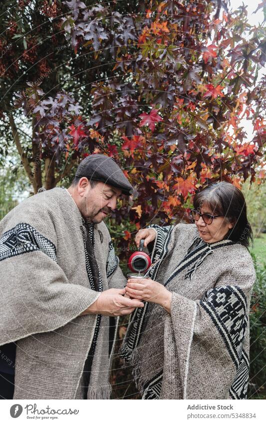 Serious mature couple in traditional Mapuche clothes with thermos and mug pouring hot yerba mate while standing in autumn garden in Temuco drink mapuche park