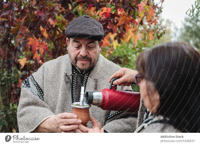 Anonymous female serving hot yerba mate to a serious male while standing in autumn garden in Temuco couple drink content thermos mapuche park together cheerful