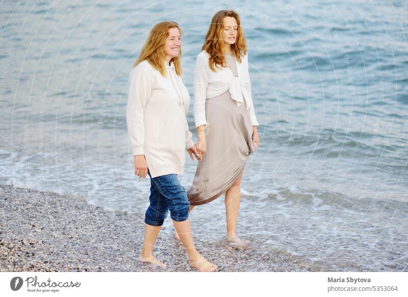 Redhead senior mother and her adult beautiful pregnant daughter are walking together on the sea shore. people maternity family look alike talk parent hang out