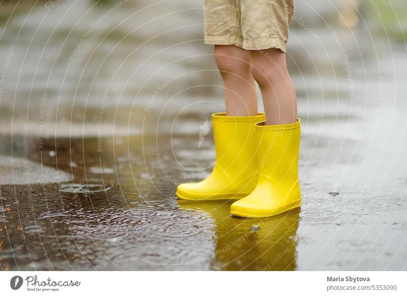Little boy wearing yellow rubber boots walking on rainy summer day in small town. Child having fun. Outdoors games for children in rain. gumboots puddle jump