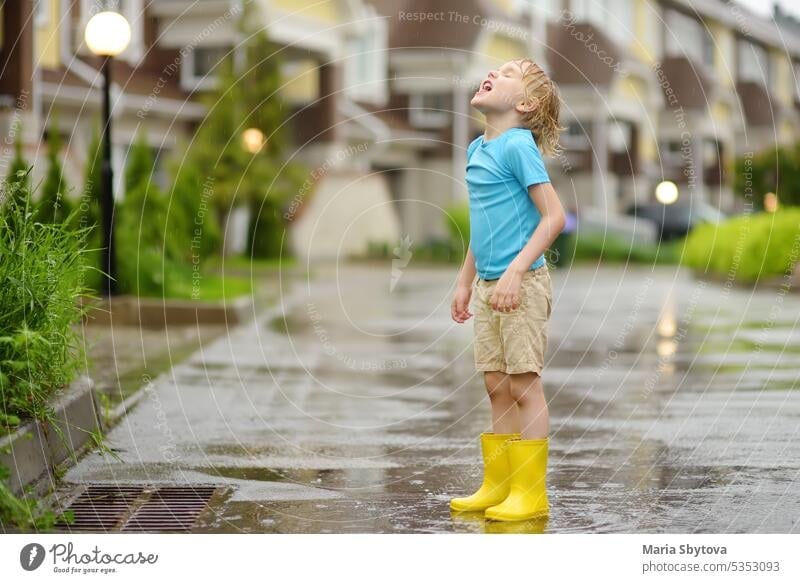 Little boy wearing yellow rubber boots walking on rainy summer day in small town. Child having fun. Kid catches raindrops with tongue. Outdoors games for children in rain.