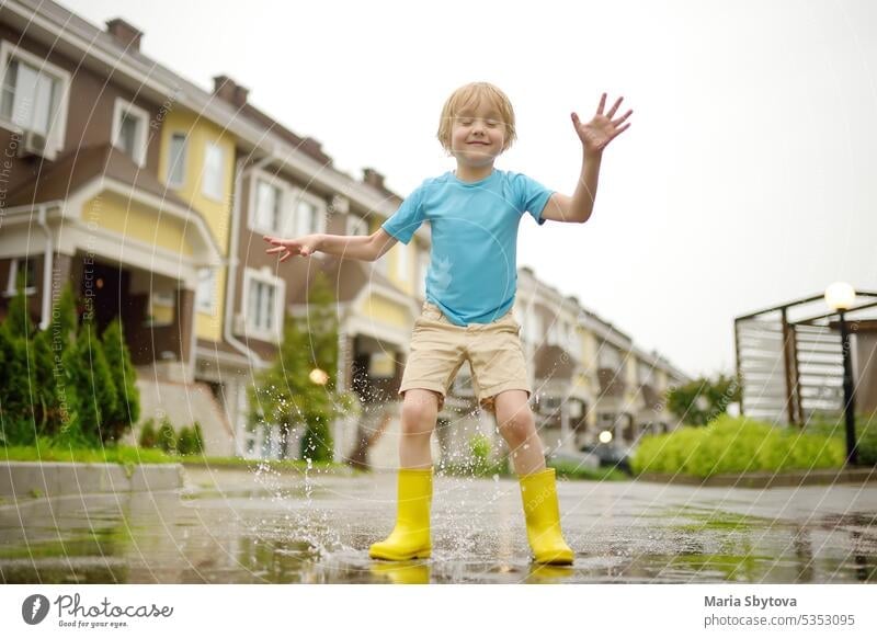 Little boy wearing yellow rubber boots jumping in puddle of water on rainy summer day in small town. Child having fun. Outdoors games for children in rain.