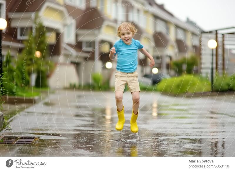 Little boy wearing yellow rubber boots jumping in puddle of water on rainy summer day in small town. Child having fun. Outdoors games for children in rain.